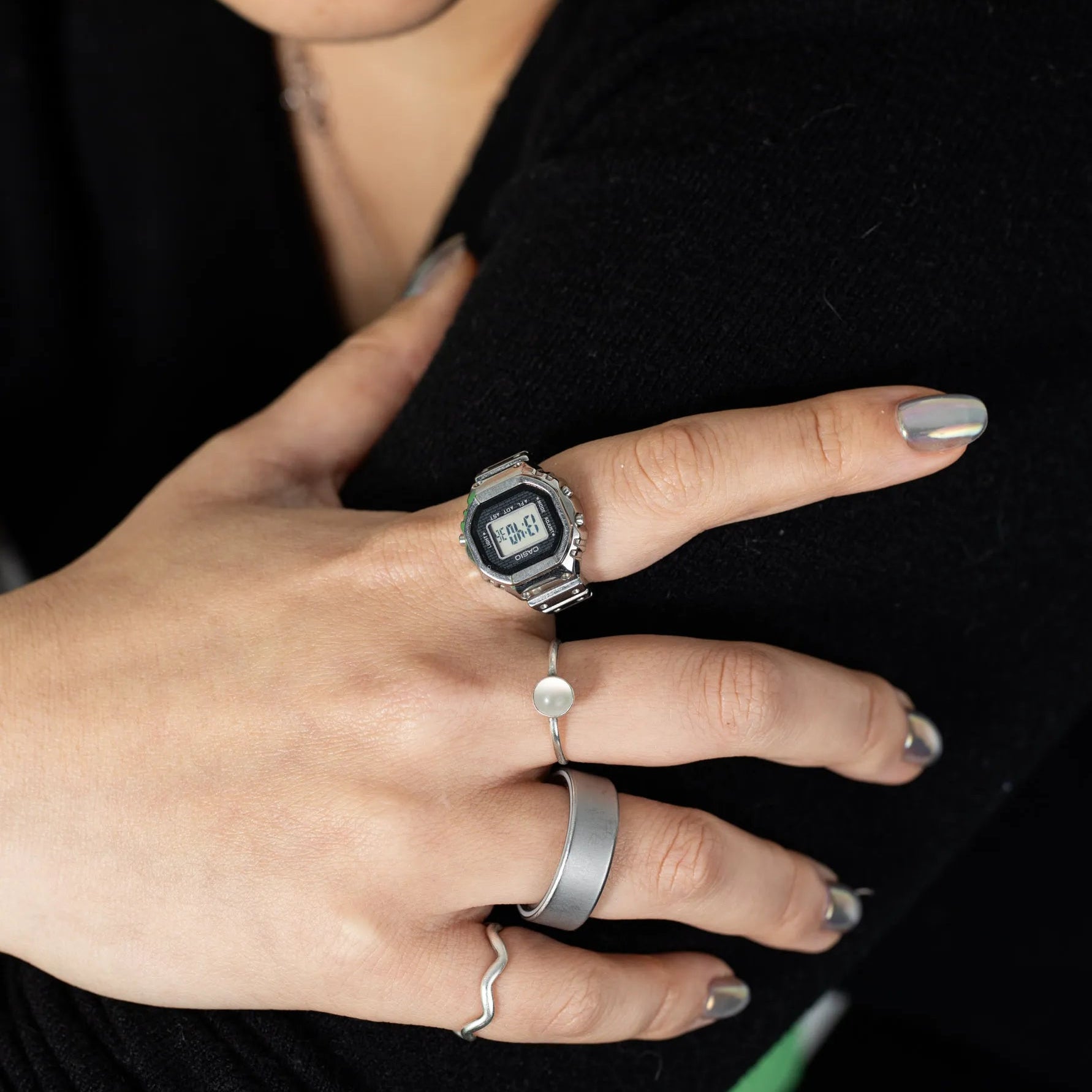 A close-up of a person's hand wearing multiple silver rings, including a watch-style ring, a slim band, a wide flat band, and a wavy chain ring, against a black top background.