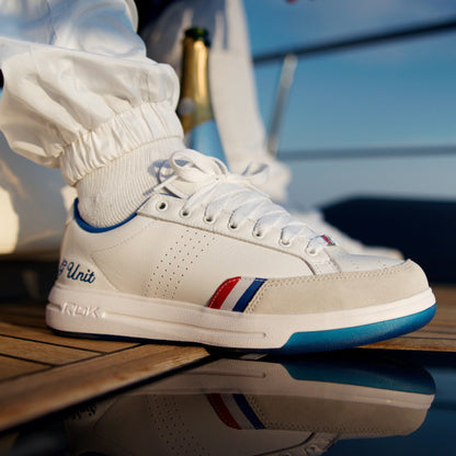 A close-up of a white low-top sneaker with blue and red accents, worn with white socks on a wooden deck with water reflection.