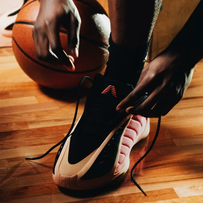 Person tying shoelaces on a black and tan sneaker with a basketball in the background on a wooden floor.