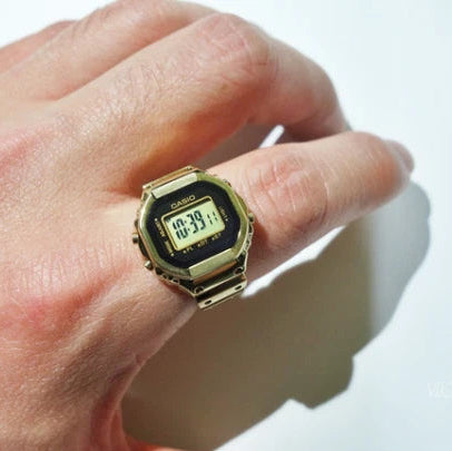 A gold-tone digital watch with a black face and green LCD display, worn on a person's ring finger against a plain white background.