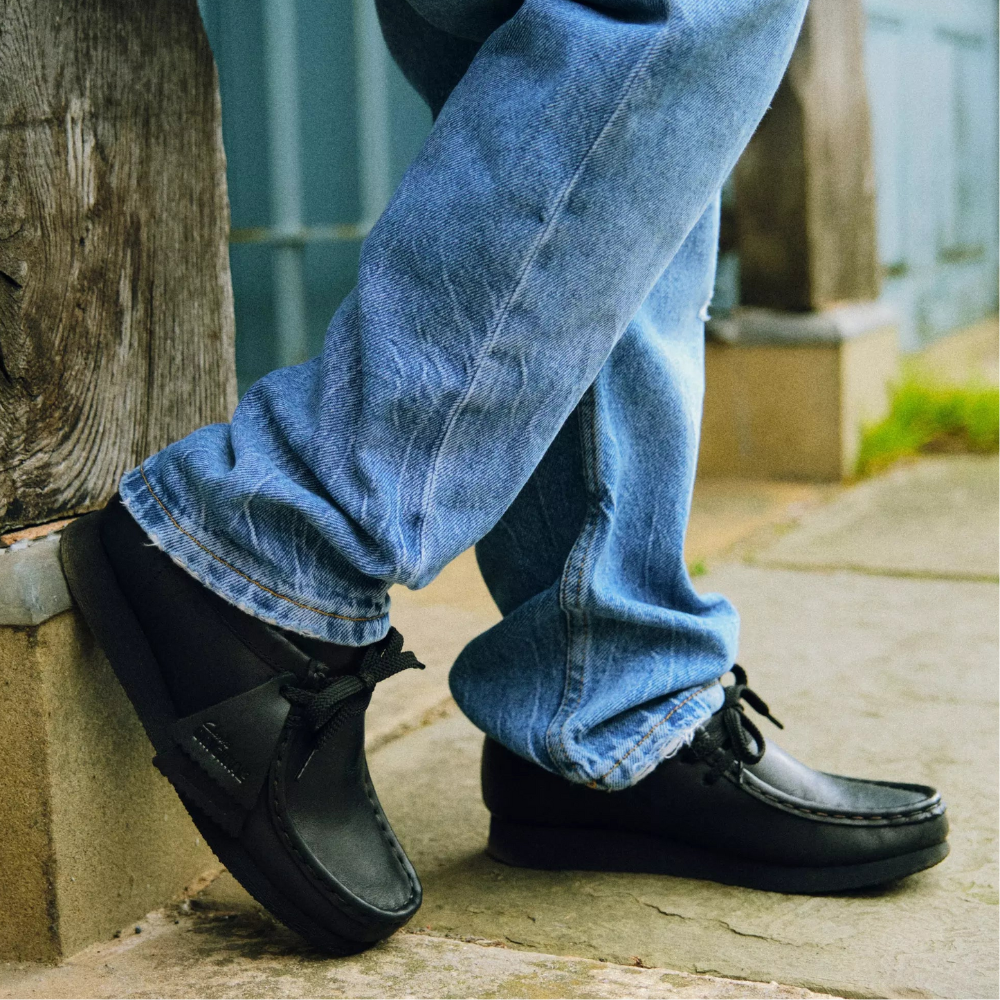 A person wearing blue jeans and black slip-on shoes, standing outdoors on a stone surface next to a wooden post.