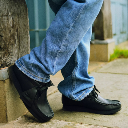 A person wearing blue jeans and black slip-on shoes, standing outdoors on a stone surface next to a wooden post.