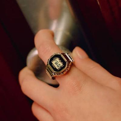 A close-up of a hand wearing a silver-toned metal ring with an integrated black digital watch face and a small screen.