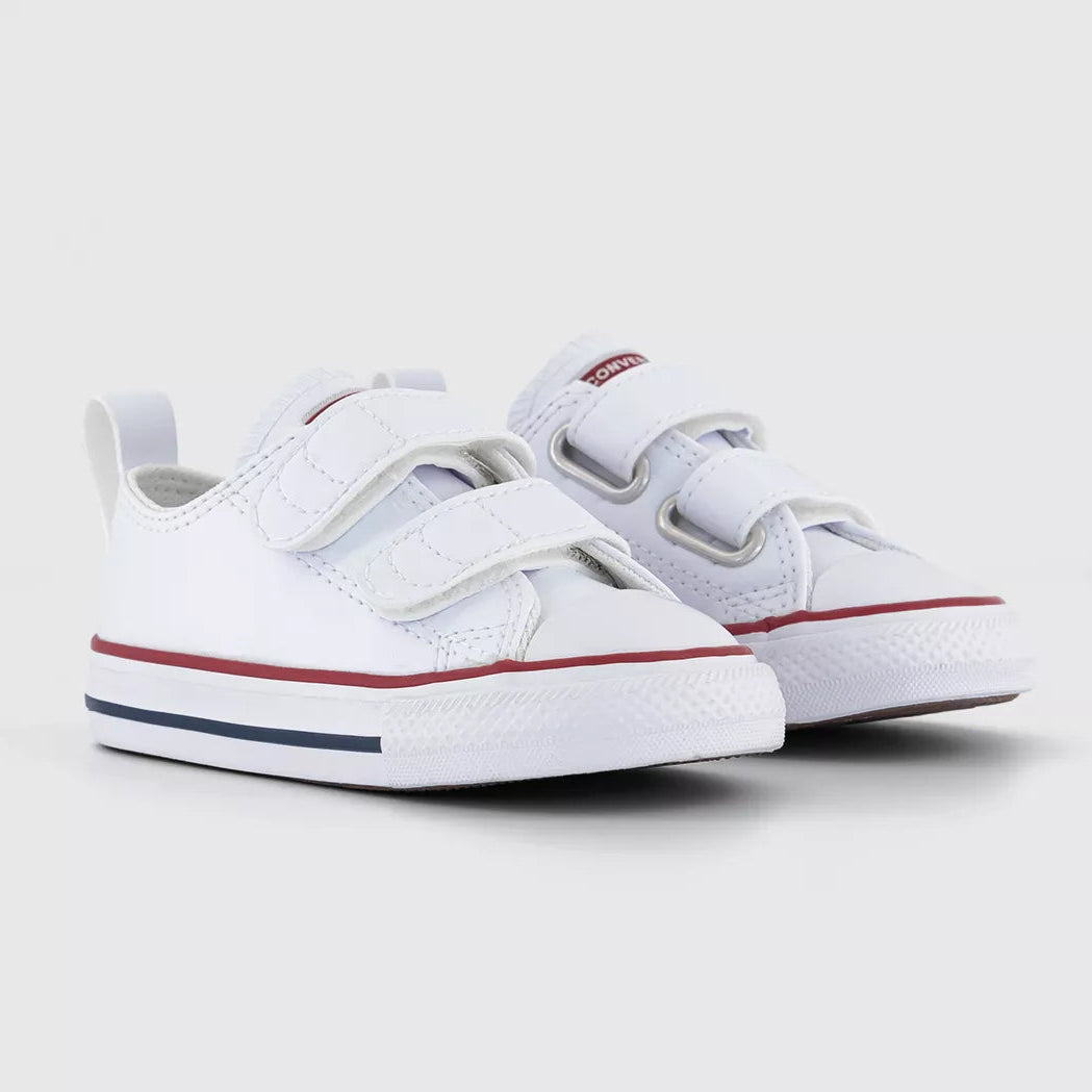A pair of white toddler low-top sneakers with hook-and-loop straps, featuring a red stripe and a navy blue stripe on the white rubber sole, set against a light gray background.